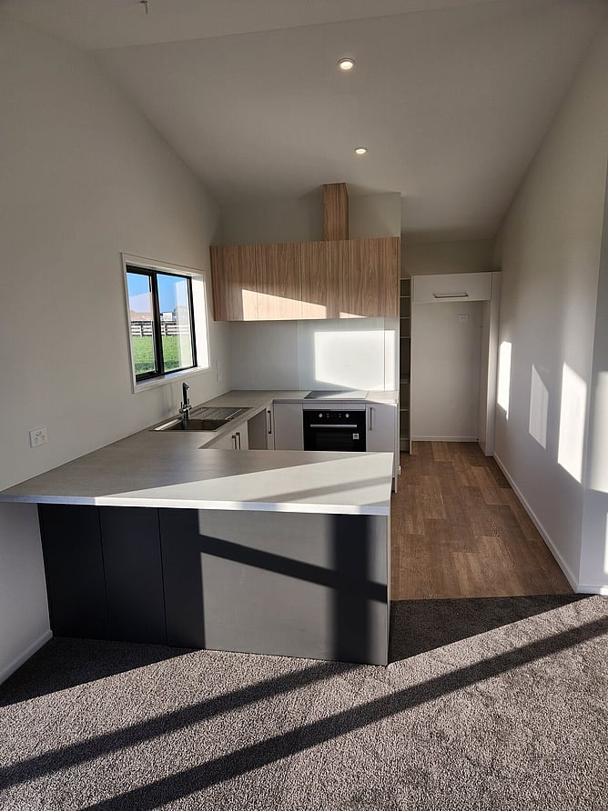 The kitchen from the living area, with a dark-fronted bench, timber overheads and the pantry beyond