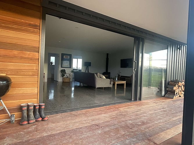 The horizontal cedar wall and Kwila deck, with the slider open to the polished concrete living area and a row of gumboots at the door