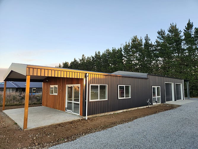 The finished Darfield showhome at dusk: black steel cladding, cedar around the entry, a timber-posted covered patio and two roller doors to the garage