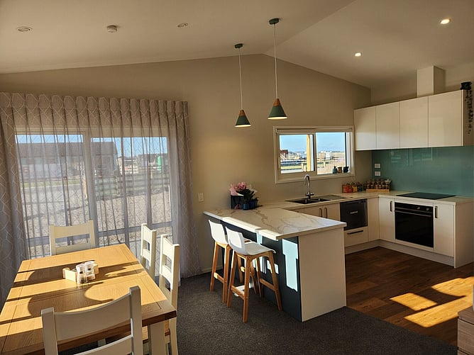 Kitchen and dining in the Darfield showhome under the raking ceiling, with sheer curtains over the slider to the covered patio