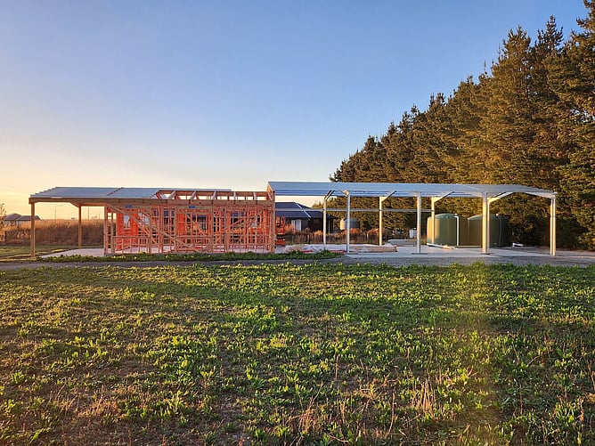 Early days on site at sunset: timber framing for the living area under the patio posts, steel portal frames for the garage bays and the water tanks alongside