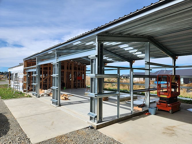 Roof on: steel portal frames for the garage and carport beside the wrapped timber framing of the living area, with a scissor lift inside