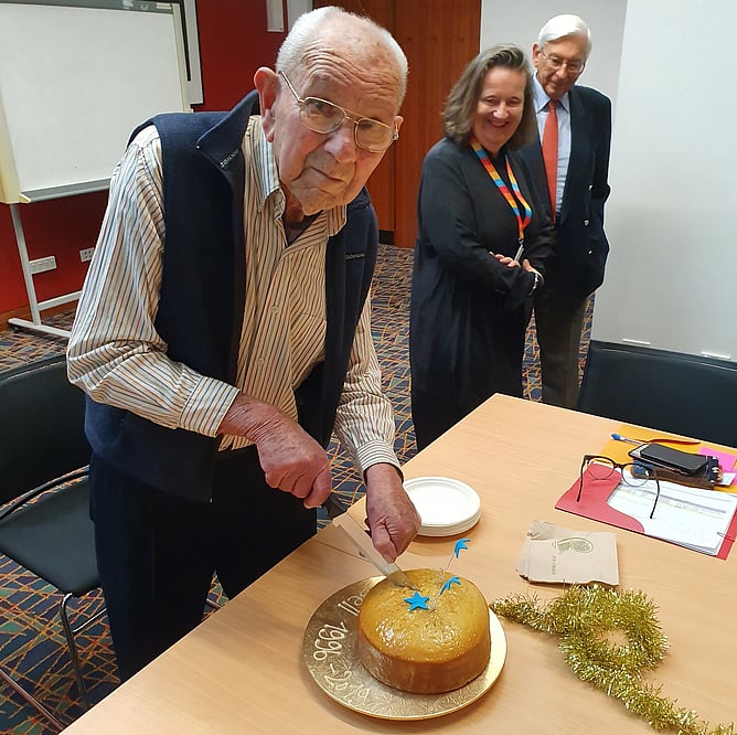 Russell Stone cutting a cake. The board reads 'Russell 1996-2019.' Russell is looking at the camera. Behind him Jane Wild and Colin David are smiling.