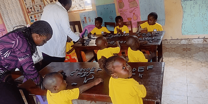 A teacher and children in bright orange uniforms participating in a group prayer activity inside a classroom at Global Star School Kakamega, Kenya.