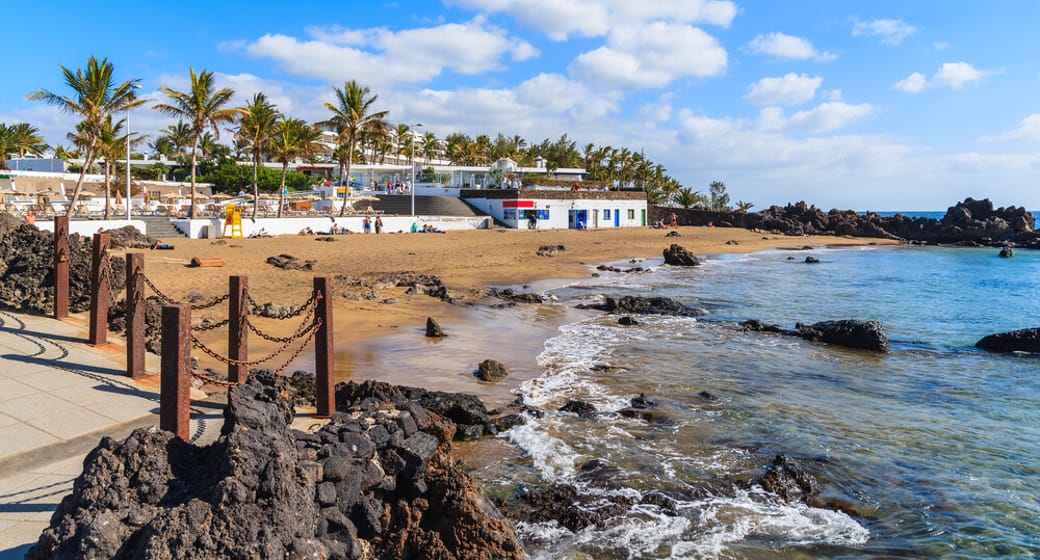 Golden sand beach with black volcanic rocks and a palm-lined promenade in Puerto del Carmen, Lanzarote