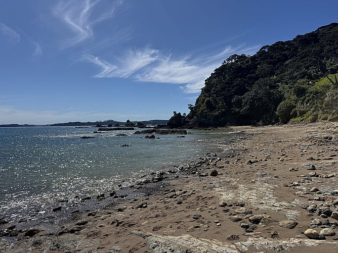Scenic view of the rocky sand shoreline at Tapeka Point Beach bordered by clear ocean waters and a steep bush-covered headland cliff